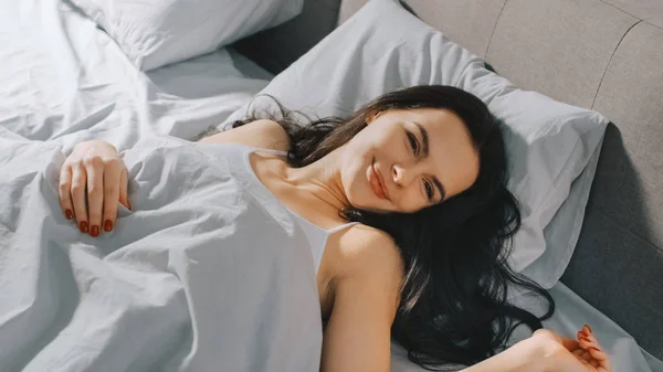 Beautiful Brunette Slowly Waking up in the Morning, Lying in a Bed, Smiles Charmingly. Sun Shines on a Happy Young Girl From the Big Window. — Stock Photo, Image