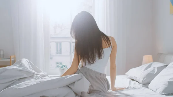 Όμορφη μελαχρινή ξυπνάει το πρωί, τεντώνει στο κρεβάτι, ο ήλιος λάμπει πάνω της από το μεγάλο παράθυρο. Ευτυχισμένο νεαρό κορίτσι χαιρετά νέα ημέρα. — Φωτογραφία Αρχείου