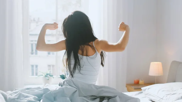 Die schöne Brünette wacht morgens auf, breitet sich im Bett aus, die Sonne scheint aus dem großen Fenster auf sie. glückliches junges Mädchen begrüßt neuen Tag. — Stockfoto