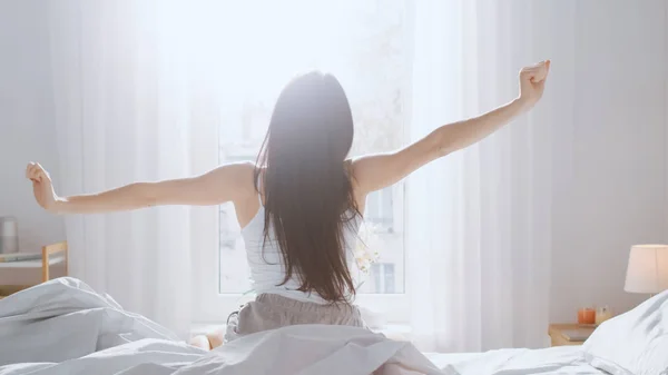 Hermosa morena se despierta por la mañana, se estira en la cama, el sol brilla en ella desde la gran ventana. Feliz joven chica saluda nuevo día . —  Fotos de Stock