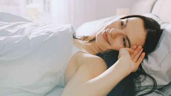 Красивая брюнетка медленно просыпается утром, лежа в постели, очаровательно улыбается. Солнце светит счастливой молодой девушке из большого окна
.