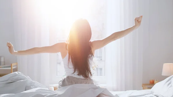 Mooie Brunette wordt wakker in de ochtend, strekt zich uit in het bed, zon schijnt op haar vanuit het grote raam. gelukkig jong meisje groet nieuwe dag met warm zonlicht flare. — Stockfoto