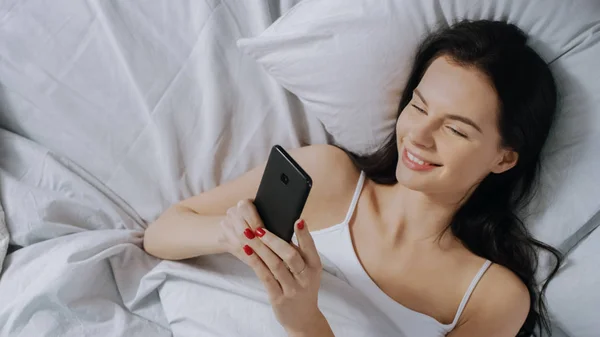 Portrait of Beautiful Brunette Using Touchscreen Smartphone in Her Bed, Chatting with Friends, Using Social Media, Posting Pictures, Sharing Thoughts. Top Down Shot.