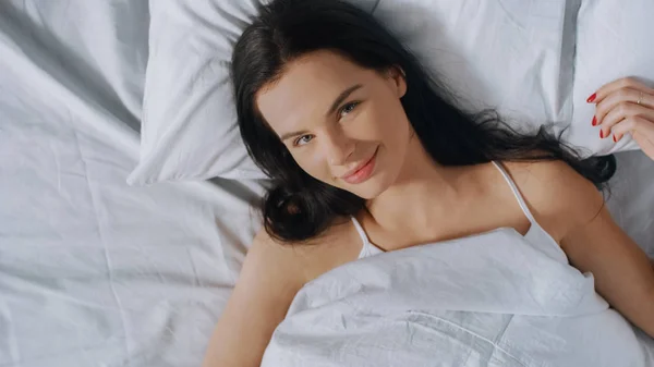 Retrato de la hermosa joven morena adorablemente acostada en su cama y sonriendo sensualmente en la cámara, su rostro está iluminado por el cálido sol de la mañana. Dulce y cálido. Top Down Shot . —  Fotos de Stock