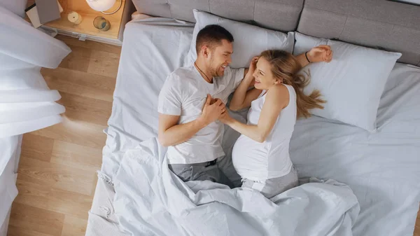 幸福的年轻夫妇一起在床上,年轻女子是怀孕和爱的伴侣触摸和爱她的肚皮温柔。顶视图拍摄. — 图库照片