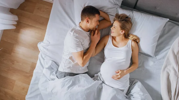 Happy Young Couple Cuddling Together in the Bed, Young Woman is Pregnant and Loving Husband Touches and Caresses Her Belly Tenderly, They Kiss. Top Down Shot.