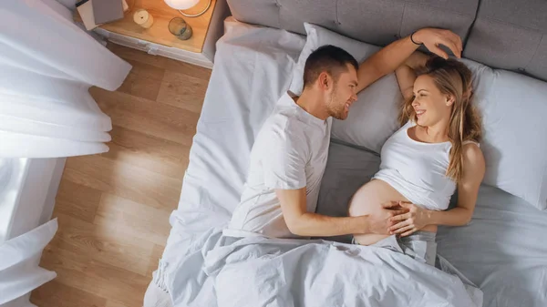 Happy Young Couple Cuddling Together in the Bed, Young Woman is Pregnant and Loving Future Father Touches and Caresses Her Belly Tenderly. Top Down Shot.