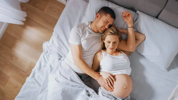 Szczęśliwy młody para cuddling razem w w łóżko, młody kobieta jest w ciąży i kochający partner dotyka i pieszczoty jej brzuch Tenderly. Słońce świeci. Strzał z góry na dół — Zdjęcie stockowe