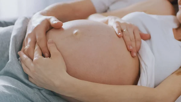 행복한 젊은 부부는 침대에서 함께 껴안고, 젊은 여성은 임신과 사랑하는 남편이 부드럽게 그녀의 배를 만지고 애무합니다. 배꼽에 카메라 클로즈업 포커스. — 스톡 사진