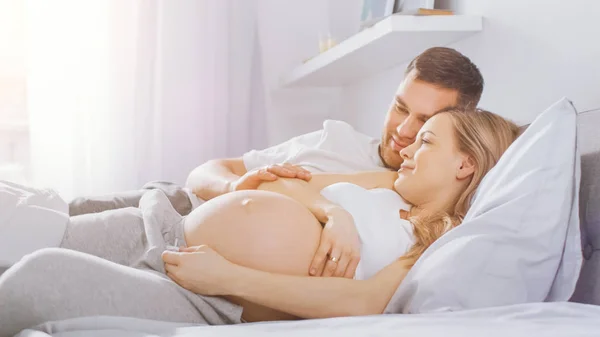 Happy Young Couple Cuddling Together in the Bed, Young Woman is Pregnant and Loving Husband Touches and Caresses Her Belly Tenderly. Camera Focus on the Belly. Warm Sunlight Enters the Room. — Stock Photo, Image