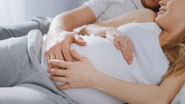 Happy Young Couple Cuddling Together in the Bed, Young Woman is Pregnant and Loving Husband Touches and Carresses Her Belly Tenderly. Foco da câmera na barriga . — Fotografia de Stock