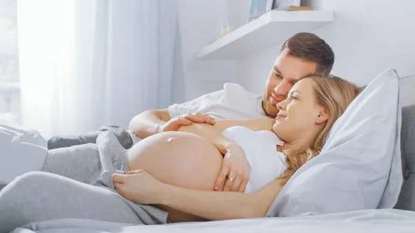 Szczęśliwa Młoda para cuddling razem w łóżku, młoda kobieta jest w ciąży i kochający mąż dotyka i pieszczoty jej brzuch Tenderly. Ostrość kamery na brzuchu. — Zdjęcie stockowe