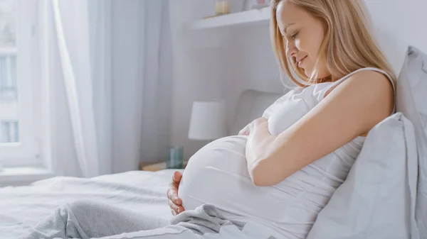 Glückliche junge schwangere Frau, die morgens im Bett liegt, sie berührt und streichelt ihren Bauch zärtlich und liebt ihr zukünftiges Baby und jeden Moment ihres Lebens. — Stockfoto