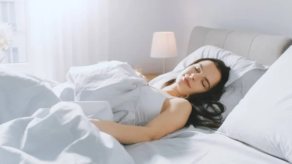 Привлекательная Брюнетка уютно спит в своей постели, в то время как ранним утром солнечные лучи освещают ее. Теплая, уютная и сладкая картина спящей красавицы — стоковое фото
