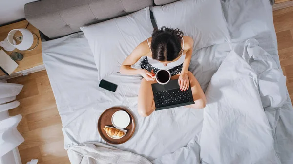 Eficiente joven niña milenaria sentada en una cama por la mañana, utiliza ordenador portátil y come cruasanes y bebidas café para el desayuno. Top Down Shot . —  Fotos de Stock