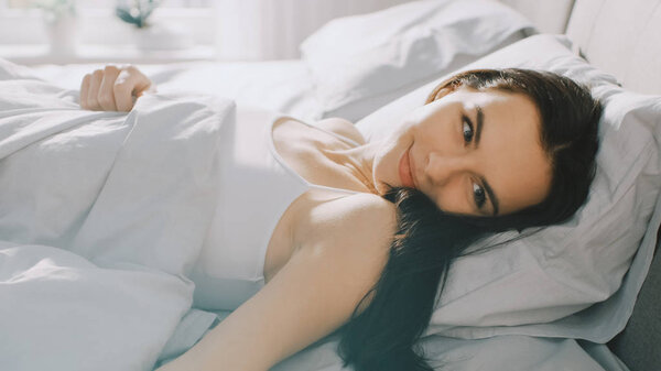 Брюнетка, лежа по утрам на кровати, завороженно смотрит в глаза и улыбается. Солнце светит счастливой молодой девушке из большого окна
.