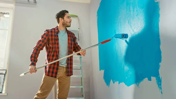 Mann in brauner Jeans und rot kariertem Hemd streicht hellblaue Farbe auf eine Walze mit langem Stiel und beginnt, eine Wand zu streichen. Farbe der Farbe ist hellblau. Renovierungen zu Hause. — Stockfoto