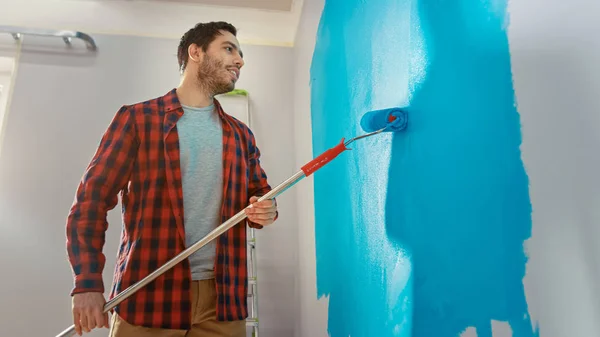Man in Brown Jeans and Red Checked Shirt está pintando una pared con un rodillo. El color de la pintura es azul claro. Renovaciones de habitaciones en casa . — Foto de Stock