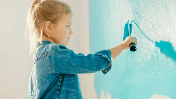 Adorable niña pequeña en Jeans Coat está pintando una pared. Ella pinta con rodillo que está cubierto de pintura azul claro. Renovaciones de habitaciones en casa . — Foto de Stock