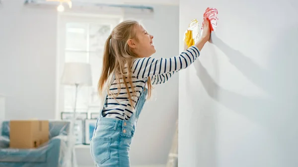 Happy Little Girl mergulha suas mãos em tinta amarela e vermelha e começa a pintar na parede. Ela está se divertindo e rindo. Casa está sendo renovada . — Fotografia de Stock