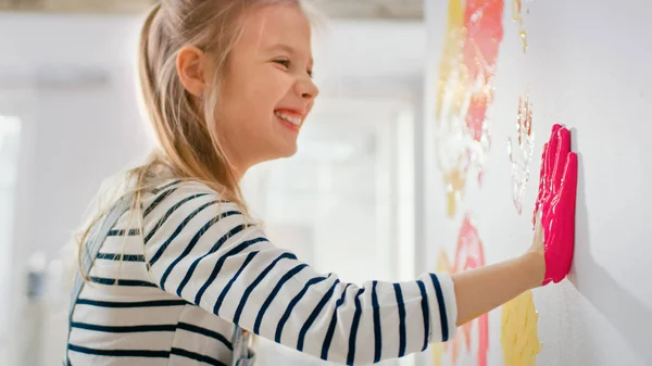 Szczęśliwa dziewczynka z rękami zanurza się w żółtym i różowym farby stawia kolorowy Handprint na ścianie. Ona jest posiadanie zabawa i śmiech. Dom jest odnowiony. — Zdjęcie stockowe