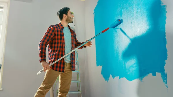 Mann in brauner Jeans und rot kariertem Hemd streicht hellblaue Farbe auf eine Walze mit langem Stiel und beginnt, eine Wand zu streichen. Farbe der Farbe ist hellblau. Renovierungen zu Hause. — Stockfoto