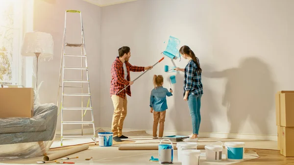 Die schöne junge Familie zeigt ihrer entzückenden kleinen Tochter, wie man Wände bemalt. Sie malen mit Rollen, die mit hellblauer Farbe überzogen sind. Schuss mit Sonnenfackel im renovierten Zimmer zu Hause. — Stockfoto