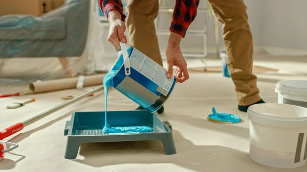 Young Man in Brown Jeans and Red Checked Shirt Picks Up a Can of Light Blue Paint and Pours it Into a Tray. Room Renovations at Home.