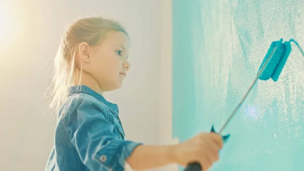 Αξιολάτρευτο μικρό κορίτσι σε τζιν παλτό ζωγραφίζει ένα τείχος. Ζωγραφίζει με ρολό που είναι καλυμμένο με μπλε χρώμα. Ανακαινίσεις δωματίων στο σπίτι. — Φωτογραφία Αρχείου