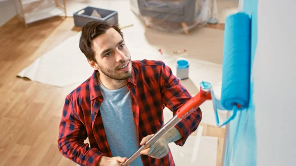 O homem de calças de ganga castanhas e camisa vermelha está a pintar uma parede com um rolo. A cor da pintura é azul claro. Renovações de quarto em casa. Tiro de cima . — Fotografia de Stock