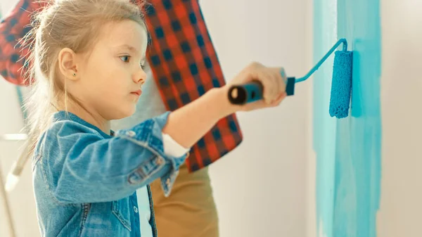 Adorable niña pequeña en Jeans Coat está pintando una pared. Ella pinta con rodillo que está cubierto de pintura azul claro. Padre pinta junto con su hija. Renovaciones de habitaciones en casa . — Foto de Stock