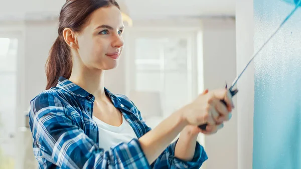 Porträtaufnahme einer jungen schönen Frau, die zu Hause eine Wand bemalt. Sie verwendet hellblaue Farbe und arbeitet mit einer Walze. Wohnungsrenovierung. — Stockfoto