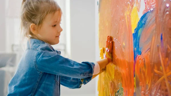 Szczęśliwa dziewczynka z rękami zanurza się w żywe farby rysuje kolorowe abstrakcje na ścianie. Ona jest posiadanie zabawa i śmiech. Dom jest odnowiony. — Zdjęcie stockowe