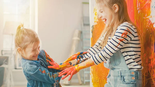 Dos pequeñas hermanas divertidas juegan y tontean con sus manos sumergidas en pintura colorida. Son felices y ríen. Objetivos de Hermandad. Redecoración en casa . —  Fotos de Stock