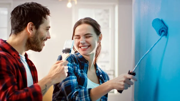 Joven hermosa pareja decorar su nuevo apartamento y tontos alrededor. Esposo y esposa están pintando la pared con rodillos que se sumergen en pintura azul claro. Son felices y se divierten. Renovaciones en casa . —  Fotos de Stock