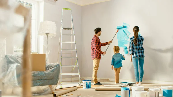 Vacker ung familj visar hur man målar väggar till sin bedårande lilla dotter. De målar med rullar som är täckta av ljusblå färg. Renovering av rum hemma. — Stockfoto