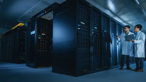 In het Data Center: twee IT-specialisten die witte Labjassen dragen, hebben discussie over het werken met server racks. Optimaliseren van het werk van de database. Cloud Computing, kunstmatige intelligentie netwerken — Stockfoto