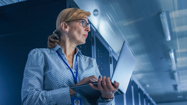 In Data Center: Low-Angle Portrait Shot of a Female IT Technician Running Maintenance Programme on Laptop, Controls Operational Server Rack. Modern High-Tech Telecommunications Operational Data Center