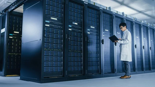 In Data Center: man it specialist dragen witte vacht staat naast server rack, maakt gebruik van laptop computer om onderhoud diagnostische hulpmiddelen uit te voeren, besturingselementen, zodat de database werkt op optimale niveau. — Stockfoto