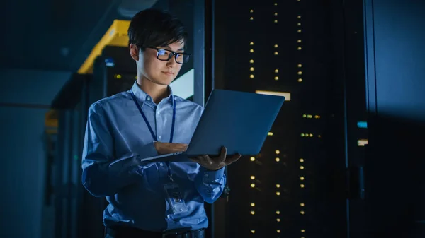 In Dark Data Center: man it specialist staat naast de rij van operationele server racks, maakt gebruik van laptop voor onderhoud. Concept voor Cloud Computing, kunstmatige intelligentie, supercomputer, cyberbeveiliging. Neon lichten — Stockfoto