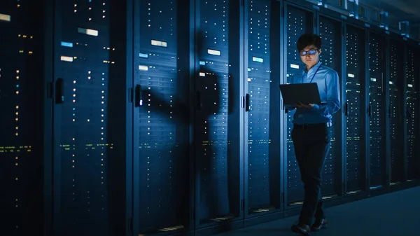 Im dunklen Rechenzentrum: männlicher IT-Spezialist läuft entlang der Reihe operativer Serverschränke, nutzt Laptop zur Wartung. Konzept für Cloud Computing, künstliche Intelligenz, Supercomputer, Cybersicherheit — Stockfoto