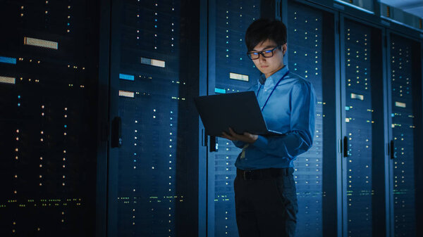In Dark Data Center: Male IT Specialist Walks along the Row of Operational Server Racks, Uses Laptop for Maintenance. Concept for Cloud Computing, Artificial Intelligence, Supercomputer, Cybersecurity