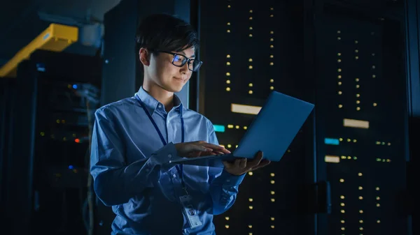 In Dark Data Center: man it specialist staat naast de rij van operationele server racks, maakt gebruik van laptop voor onderhoud. Concept voor Cloud Computing, kunstmatige intelligentie, supercomputer, cyberbeveiliging. Neon lichten — Stockfoto