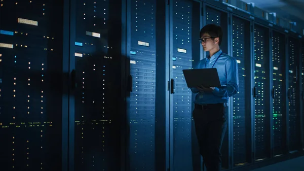 Im dunklen Rechenzentrum: männlicher IT-Spezialist läuft entlang der Reihe operativer Serverschränke, nutzt Laptop zur Wartung. Konzept für Cloud Computing, künstliche Intelligenz, Supercomputer, Cybersicherheit — Stockfoto