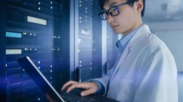 W centrum danych: mężczyzna It Specialist noszenie White Coat stoi obok rack Server, używa komputera przenośnego do uruchomienia narzędzia Diagnostyka konserwacji, kontroluje, aby baza danych pracuje w optymalnym poziomie. — Zdjęcie stockowe