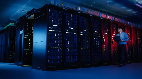 V datovém centru: samčí specialista na IT kráčí po řadě operačních stojanů na serveru, používá přenosný počítač k údržbě. Koncept pro telekomunikace, Cloud Computing, umělá inteligence, superpočítač — Stock fotografie