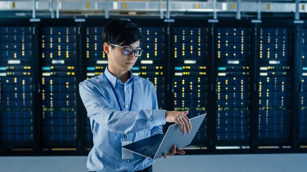 En Dark Data Center: Male IT Specialist se encuentra junto a la fila de bastidores de servidores operativos, utiliza el ordenador portátil para el mantenimiento. Concepto para Cloud Computing, Inteligencia Artificial, Supercomputadora, Ciberseguridad — Foto de Stock