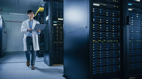 In Data Center: Male IT Technician Wearing White Coat Walking alongside Server Racks, Uses Laptop Computer to Run Maintenance Diagnostics Tools. He is Wearing Lab Coat and Working with Data.