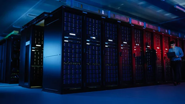 V datovém centru: samčí specialista na IT kráčí po řadě operačních stojanů na serveru, používá přenosný počítač k údržbě. Koncept pro telekomunikace, Cloud Computing, umělá inteligence, superpočítač — Stock fotografie