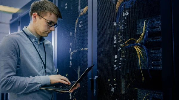 En el centro de datos, el ingeniero de TI se para antes de trabajar en el estante del servidor realizando comprobaciones y diagnósticos de mantenimiento rutinario mediante computadora portátil. Equipo de hardware de computadora visible, luces led de cables de fibra óptica de banda ancha . —  Fotos de Stock